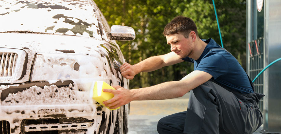 Θεματική εικόνα για το πώς να πλύνω το αυτοκίνητό μου. Νεαρός τρίβει με σφουγγάρι το μπροστινό μέρος του αμαξιού του.